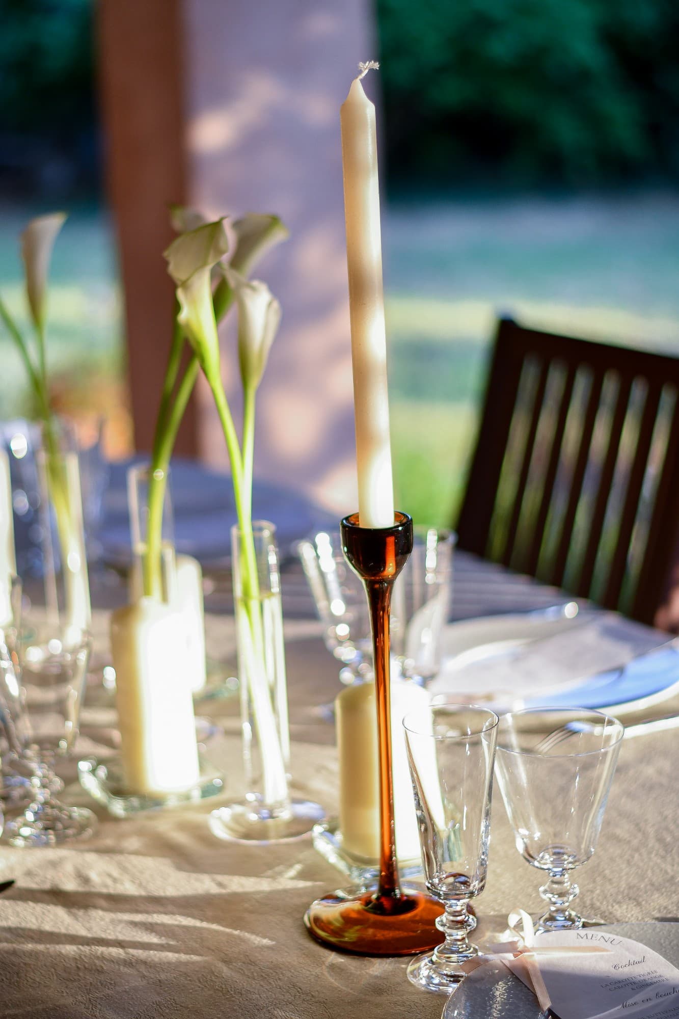 Table dressée en extérieur, bougie sur un bougeoir ambré, arums blancs et verres en cristal